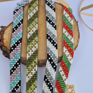May include: Four beaded bracelets with geometric patterns in various colors, including black, white, green, red, and blue. The bracelets are displayed on a wooden surface.