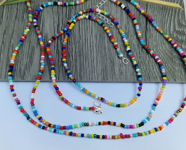 May include: A collection of colorful beaded necklaces. The necklaces feature small, round beads in a variety of colors, including red, yellow, blue, green, and white. Each necklace has a silver-colored clasp and adjustable chain. The necklaces are displayed on a gray wooden surface.