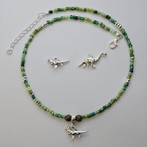 May include: A green beaded necklace with a silver dinosaur charm. The necklace is made with small, green glass beads and has a silver clasp. The dinosaur charm is a small, silver Tyrannosaurus Rex.