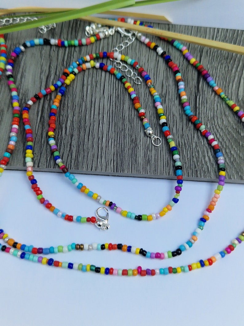 May include: A collection of colorful beaded necklaces. The necklaces feature small, round beads in a variety of colors, including red, yellow, blue, green, and purple. Each necklace has a silver-colored clasp and adjustable chain.