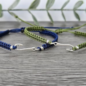 May include: Two friendship bracelets with a silver fish hook charm. One bracelet is dark blue and the other is green.