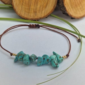 May include: A turquoise stone bracelet with a brown cord. The bracelet features several irregularly shaped turquoise stones strung together. The cord is adjustable with a sliding knot. Small gold-coloured beads are at each end of the turquoise stones.