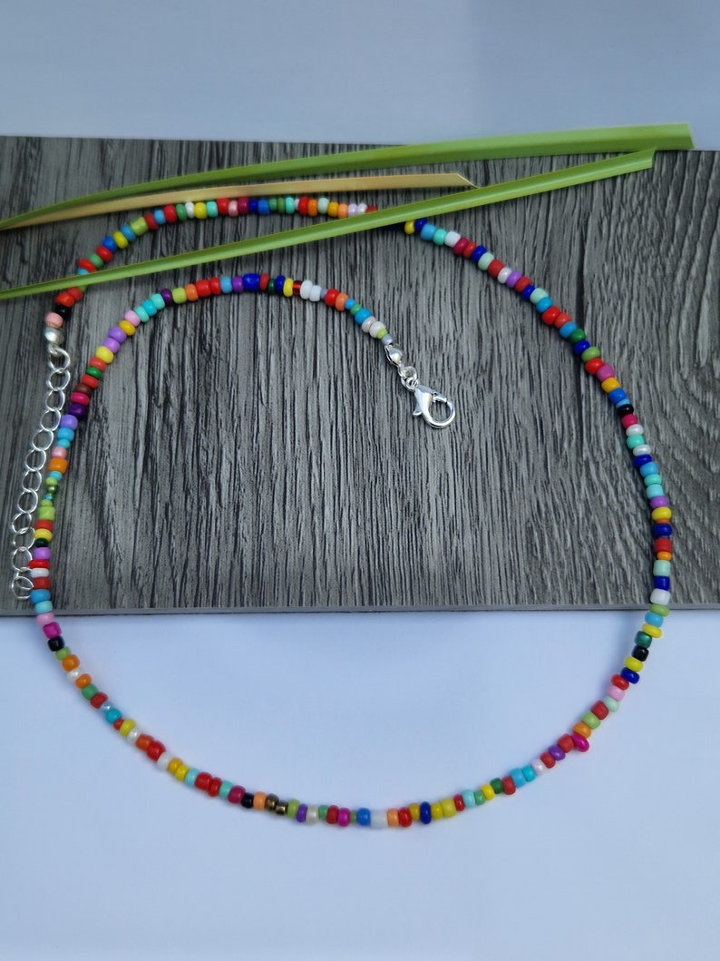 May include: A colorful beaded necklace with a silver clasp. The necklace features small, round beads in a variety of colors, including red, orange, yellow, green, blue, and purple. The necklace is displayed on a gray wooden surface.