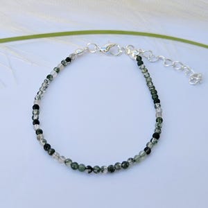 May include: A delicate bracelet made with small, round beads in shades of green, black, and clear. The bracelet has a silver clasp.