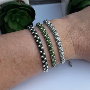 Könnte beinhalten: Drei Armbänder aus grünen und silbernen Perlen. Die Armbänder sind miteinander verwoben und haben ein schlichtes Design.