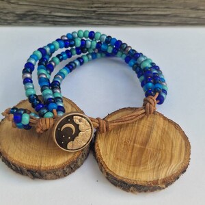 May include: Multi-strand beaded bracelet featuring blue, turquoise, and silver beads. The bracelet has a brown leather cord with a metal charm depicting a crescent moon and clouds.