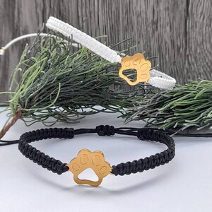May include: Two adjustable bracelets with gold paw print charms. One bracelet is black and the other is white.
