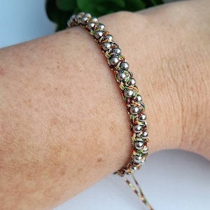 Könnte beinhalten: Ein geflochtenes Armband in Silber und verschiedenen Farben mit silbernen Perlen. Das Armband hat einen verschiebbaren Knotenverschluss.