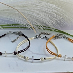 Puede incluir: Tres pulseras con un dije de cruz plateada. Las pulseras están hechas de cordón marrón, blanco y amarillo.