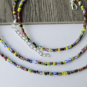 May include: Three colorful beaded necklaces with a variety of small, round beads in red, yellow, blue, green, and black. The necklaces have silver-colored clasps and extenders. The background is a light wood grain.