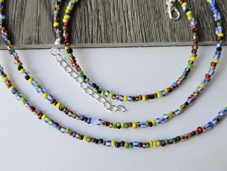 May include: Three colorful beaded necklaces with a variety of small, round beads in red, yellow, blue, green, and black. The necklaces have silver-colored clasps and extenders. The background is a light wood grain.