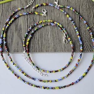 May include: Three colorful beaded necklaces with silver clasps. The necklaces feature small, round beads in various colors, including red, yellow, blue, green, and black. The necklaces are arranged in a layered style on a wooden surface.