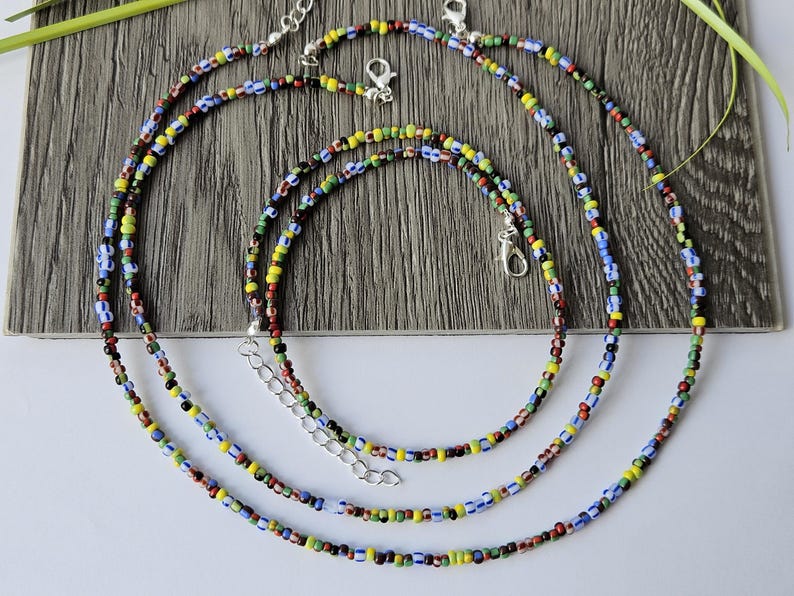 May include: Three colorful beaded necklaces with silver clasps. The necklaces feature small, round beads in various colors, including red, yellow, blue, green, and black. The necklaces are arranged in a layered style on a wooden surface.