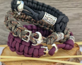 Puede incluir: Tres pulseras trenzadas de paracord con detalles metálicos plateados. La pulsera superior es negra, la pulsera del medio es de camuflaje y la pulsera inferior es burdeos.