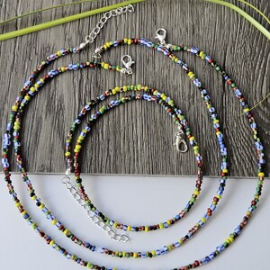 May include: Three colorful beaded necklaces with silver-toned clasps and extenders. The beads are small and feature a mix of colors including red, yellow, blue, green, and black. The necklaces are arranged on a wooden surface.