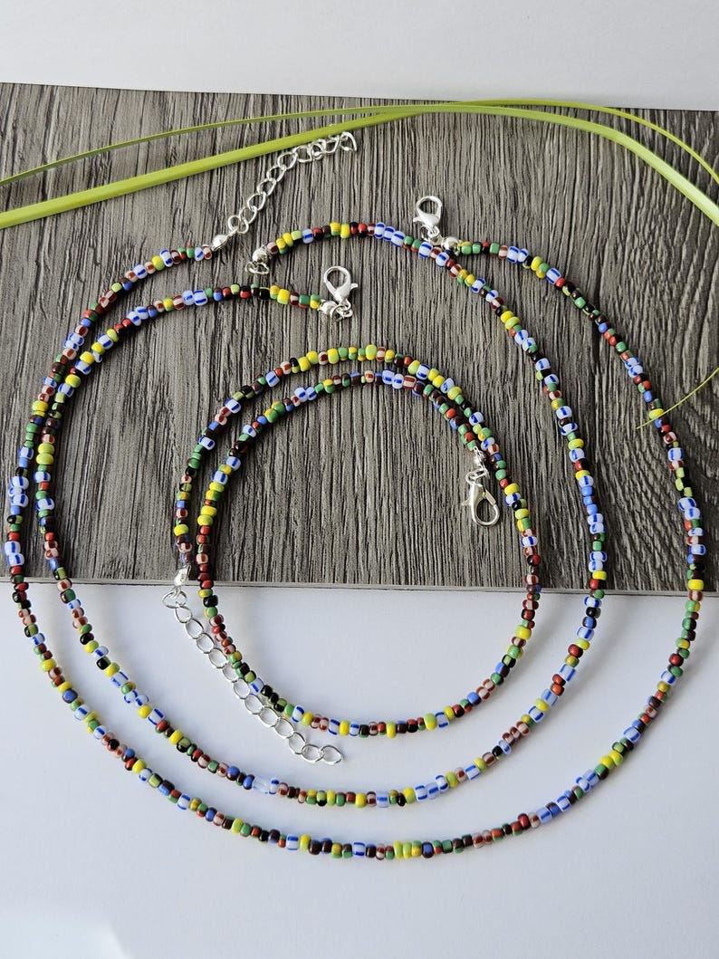 May include: Three colorful beaded necklaces with silver-toned clasps and extenders. The beads are small and feature a mix of colors including red, yellow, blue, green, and black. The necklaces are arranged on a wooden surface.