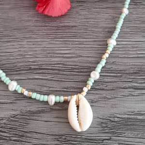 May include: A light green and white beaded necklace with a white shell pendant. The necklace is on a wooden surface.