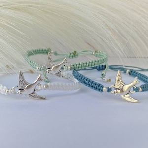 Könnte beinhalten: Drei geflochtene Armbänder mit silbernen Taubenanhängern. Die Armbänder sind in Weiß, Hellgrün und Blau.