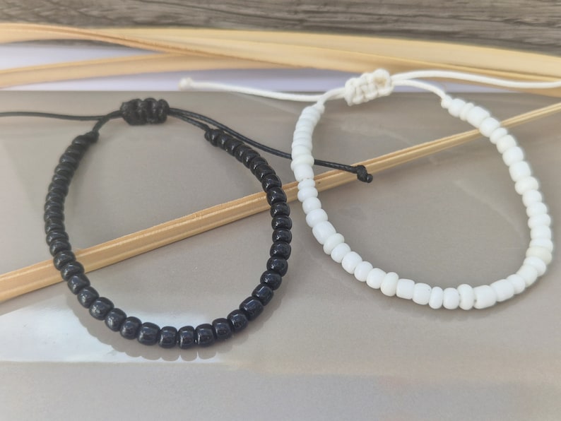 Puede incluir: Dos pulseras de cuentas, una negra y otra blanca, se muestran sobre una superficie de madera. Las pulseras est&aacute;n hechas de peque&ntilde;as cuentas redondas y est&aacute;n atadas juntas con un nudo.