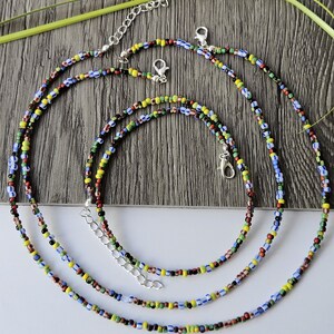 May include: Three colorful beaded necklaces with silver-toned clasps and extenders. The beads are small and feature a mix of colors including red, yellow, blue, green, and black. The necklaces are displayed on a wooden surface.