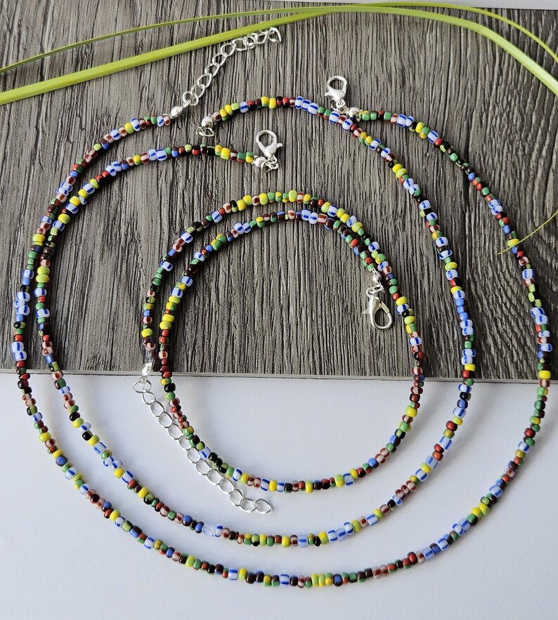 May include: Three colorful beaded necklaces with silver-toned clasps and extenders. The beads are small and feature a mix of colors including red, yellow, blue, green, and black. The necklaces are displayed on a wooden surface.