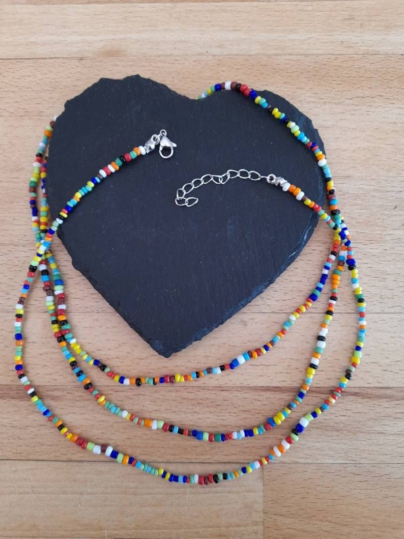 May include: A multi-strand necklace made of colorful glass beads. The necklace is layered and features a variety of colors, including red, orange, yellow, green, blue, and purple. The necklace is finished with a silver clasp.