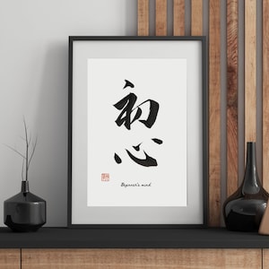 以下が含まれることがあります： 黒い額縁に入った白黒の書道アートプリント。木製の棚に置かれ、黒い花瓶が2つと金色の花瓶が1つある。書道は「初心」と書いてあり、その下に「Beginner's mind」と書いてある。