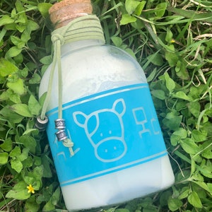 May include: A glass bottle with a cork stopper and a blue label featuring a cow illustration. The bottle is filled with a white liquid and has a green string tied around the neck.