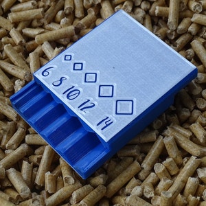 May include: A blue and white plastic container with a sliding tray, featuring the numbers 6, 8, 10, 12, and 14, along with diamond-shaped symbols. The container is resting on a bed of small, cylindrical wood pellets.