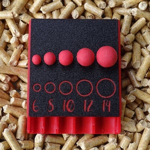 May include: A black and red tool with red spheres and circles of varying sizes. The tool has the numbers 6, 8, 10, 12, and 14 printed on it. The tool is set against a background of wood pellets.
