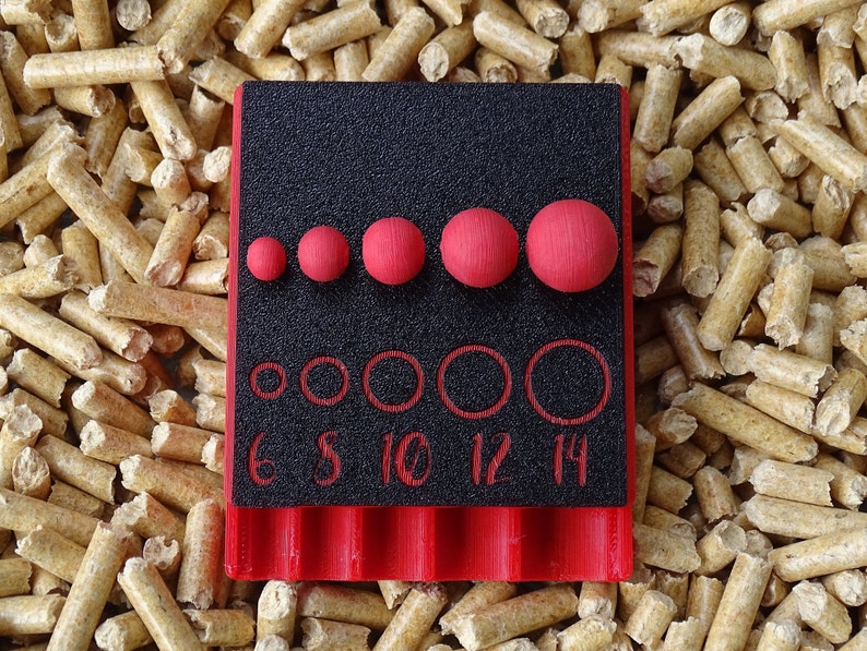 May include: A black and red tool with red spheres and circles of varying sizes. The tool has the numbers 6, 8, 10, 12, and 14 printed on it. The tool is set against a background of wood pellets.