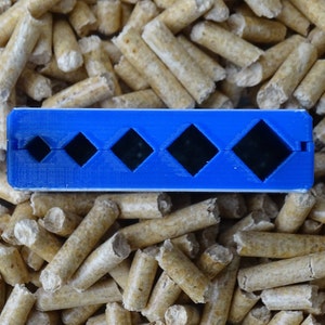 May include: A blue plastic tool with four diamond-shaped cutouts of varying sizes. The tool is resting on a bed of light brown wood pellets. The tool appears to be designed for measuring or shaping.