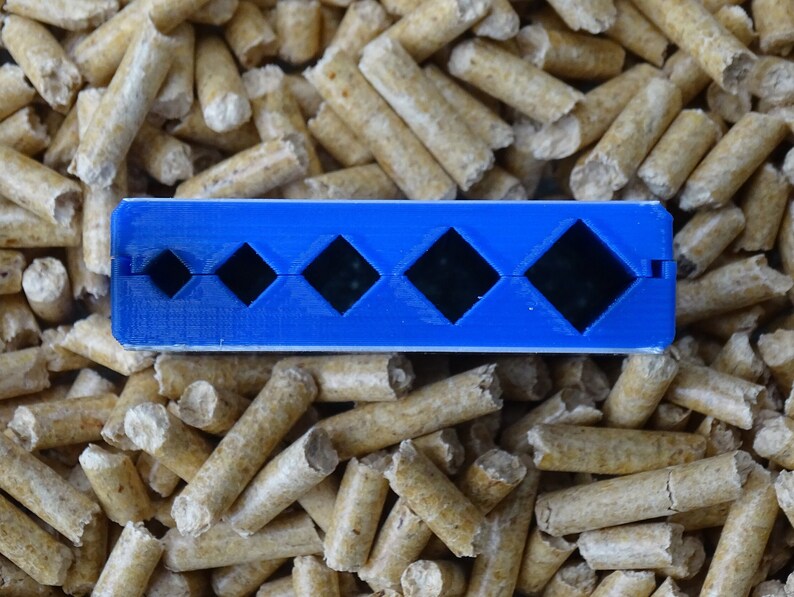 May include: A blue plastic tool with four diamond-shaped cutouts of varying sizes. The tool is resting on a bed of light brown wood pellets. The tool appears to be designed for measuring or shaping.