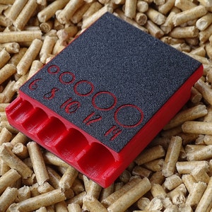 May include: A red and black tool with multiple circular openings and labeled sizes. The tool has a textured black surface with red numbers indicating sizes 6, 8, 10, 12, and 14. The tool is resting on a bed of wood pellets.