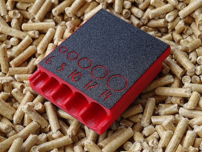 May include: A red and black tool with multiple circular openings and labeled sizes. The tool has a textured black surface with red numbers indicating sizes 6, 8, 10, 12, and 14. The tool is resting on a bed of wood pellets.