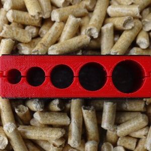 May include: A bright red, rectangular tool with five circular openings of varying sizes. The tool is set against a background of small, cylindrical, light brown wood pellets. The tool appears to be made of plastic.