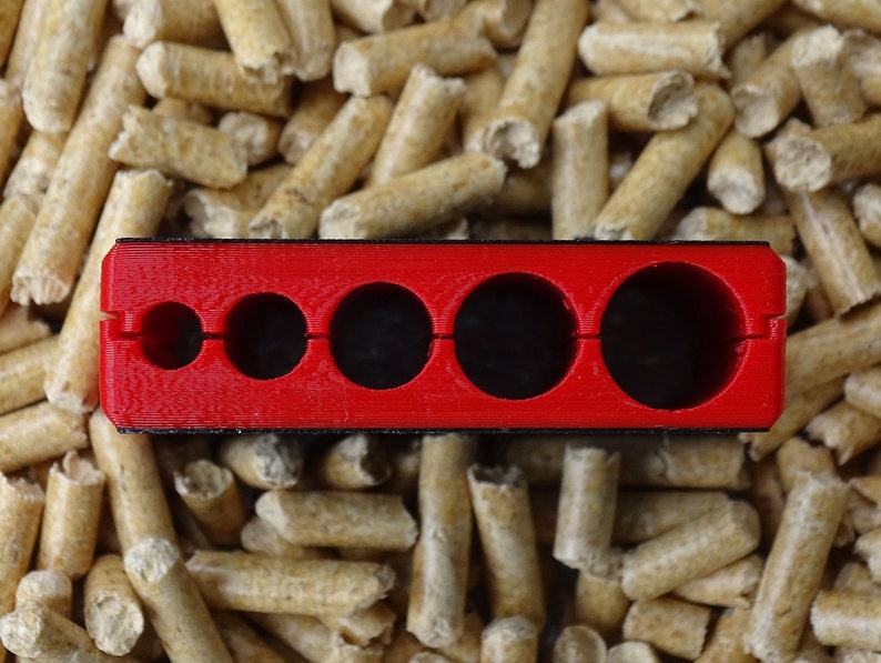 May include: A bright red, rectangular tool with five circular openings of varying sizes. The tool is set against a background of small, cylindrical, light brown wood pellets. The tool appears to be made of plastic.