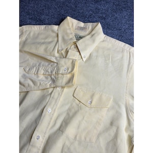 May include: A pale yellow button-down shirt with a button-down collar and a chest pocket. The long-sleeved shirt has buttoned cuffs and a label at the back of the collar. The shirt is made of a light-colored fabric.