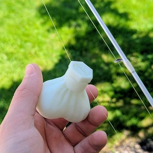 May include: A white, matte-finished, 3D-printed fishing lure shaped like a rounded, pleated pouch with a hexagonal screw-top cap. The lure features a smooth, bulbous body and is attached to a thin, clear fishing line near a silver fishing rod.