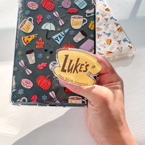 Può includere: Una custodia trasparente per tablet con sfondo scuro e icone colorate tra cui zucche, tazze da caffè con l'etichetta Luke's, ciambelle, ombrelli e un gagliardetto Yale. È fissato un supporto circolare per telefono con la scritta gialla Luke's Stars Hollow, Connecticut.