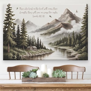 May include: A framed canvas print featuring a watercolor landscape of mountains, trees, and a river. The artwork is in shades of green, gray, and brown. Text at the top reads, "Those who trust in the Lord will renew their strength; they will soar on wings like eagles. Isaiah 40:31."