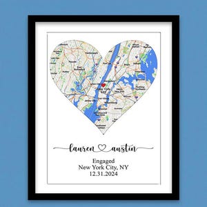 May include: A framed print featuring a heart-shaped map of New York City with a red heart icon over Manhattan. Below the map, the text reads: lauren ♡ austin, Engaged, New York City, NY, 12.31.2024. The print is enclosed in a simple black frame.