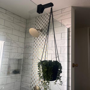 Peut inclure: Un pot de plante suspendu noir avec une plante verte retombante suspendue au plafond de la douche. Le pot est maintenu par une chaîne et un support noirs. Les murs de la douche sont carrelés avec des accents blancs et dorés.