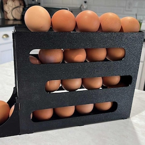 Puede incluir: Un soporte negro para huevos lleno de huevos marrones. El soporte tiene tres estantes horizontales y una base curva. Los huevos están dispuestos ordenadamente, algunos en la parte superior y otros en los estantes. El soporte está sobre una encimera blanca.