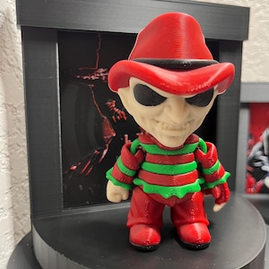 Puede incluir: Una figura impresa en 3D de un personaje que lleva un sombrero rojo, pantalones rojos y una camisa a rayas verdes y rojas. La figura tiene una cara blanca con ojos negros y una sonrisa siniestra. La figura se exhibe en una plataforma negra.