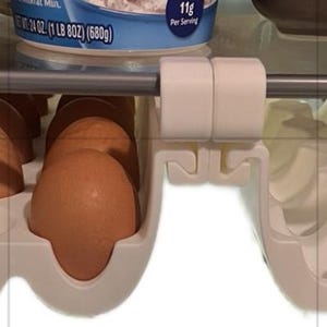 May include: A white plastic fridge egg holder with a metal bar. The egg holder is designed to hold a dozen eggs. Several brown eggs are visible in the egg holder. The egg holder is mounted on a shelf.