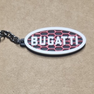 Puede incluir: Llavero Bugatti con un emblema ovalado. El emblema presenta un borde blanco, un fondo negro y un diseño de panal de abeja rojo. La palabra "BUGATTI" está en letras blancas. Incluye un llavero y una cadena plateados.