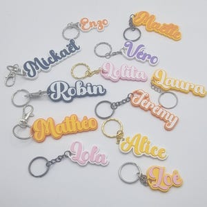 Peut inclure: Plusieurs porte-clés personnalisés avec des noms variés. Chaque porte-clé affiche un nom différent dans une police stylisée, attaché à un anneau métallique. Les noms incluent Michael, Enzo, Maelle, Vero, Lolita, Laura, et d'autres.