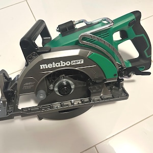 May include: A green and gray circular saw with the text "metabo HPT" on the side. The saw has a black handle and a silver blade guard. The saw is designed for cutting wood and other materials.