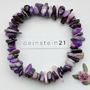 Op de afbeelding: Een armband gemaakt van gepolijste, onregelmatige paarse stenen. De stenen variëren in tinten van licht lavendel tot diep paars en zwart. De armband is cirkelvormig en ligt op een wit oppervlak. De tekst "deinstein21" is gedrukt op een wit label.